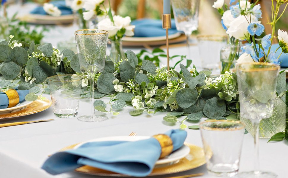 Faux eucalyptus garland with silk silver dollar leaves and plastic berries draped across a table setting with blue napkins and gold chargers.