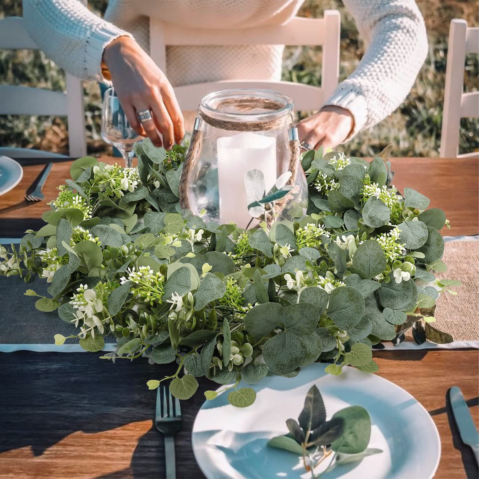 Faux eucalyptus garland with white flowers arranged around a glass jar with a candle on a wooden table setting