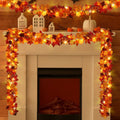Artificial maple leaf garland with LED lights draped over a fireplace mantel with decorative houses and pumpkins.