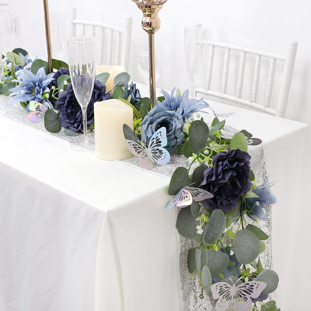 Artificial eucalyptus garland with navy blue flowers and silver butterflies draped on a white tablecloth with candles and champagne glasses.