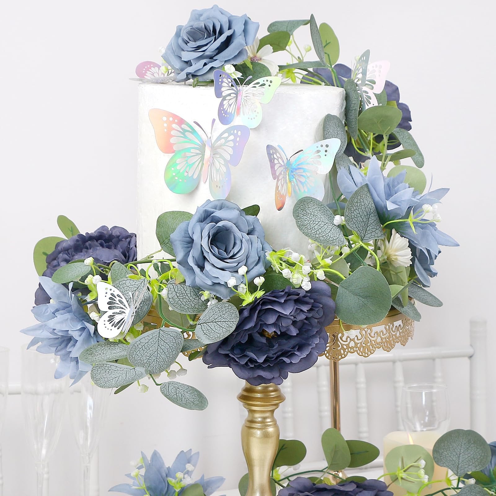 Artificial eucalyptus garland with navy blue flowers and iridescent butterflies decorating a white cake on a gold stand.