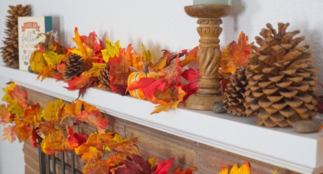 Artificial maple leaf fall garland with pinecones and a small pumpkin draped over a fireplace mantel.