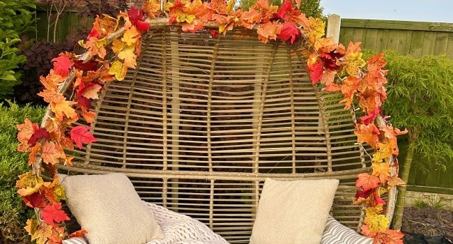Artificial maple leaf garland draped over a hanging chair with pillows and a knit blanket, set in a garden.