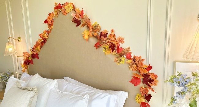 Artificial maple leaf garland draped over a headboard with pillows and a lamp.