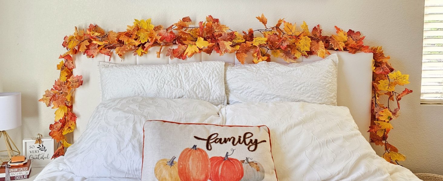 Artificial maple leaf garland draped over a headboard with white pillows and a 'family' pumpkin pillow.