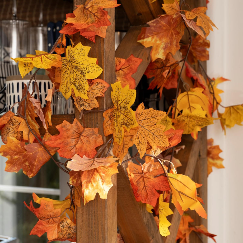 Artificial maple leaf garland with vibrant orange and yellow leaves draped on a wooden structure, showcasing autumn colors.