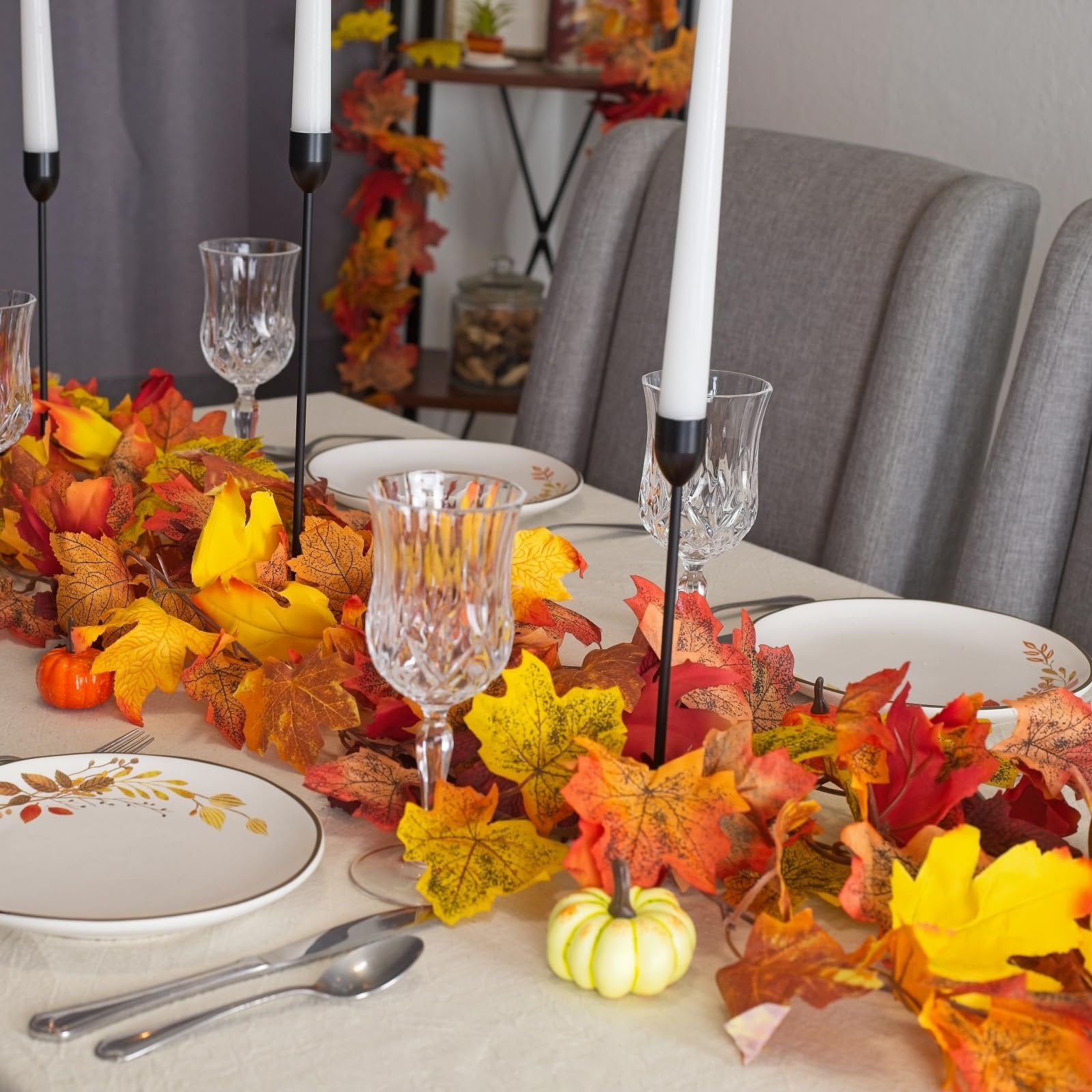 Artificial maple leaf garland draped across a dining table set for a meal, with pumpkins and candles.