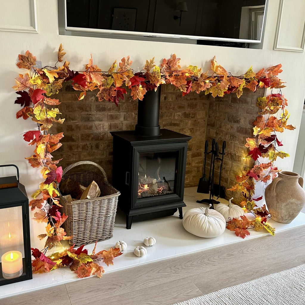 Artificial maple leaf garland with lights draped over a fireplace mantel with a wood-burning stove, basket of logs, and pumpkins.
