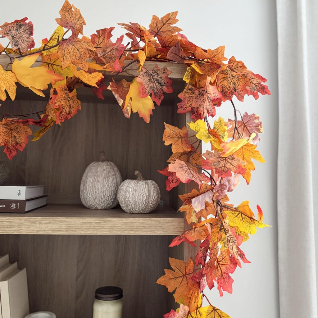 Artificial maple leaf garland with autumn colors draped over a wooden shelf with decorative pumpkins and books.