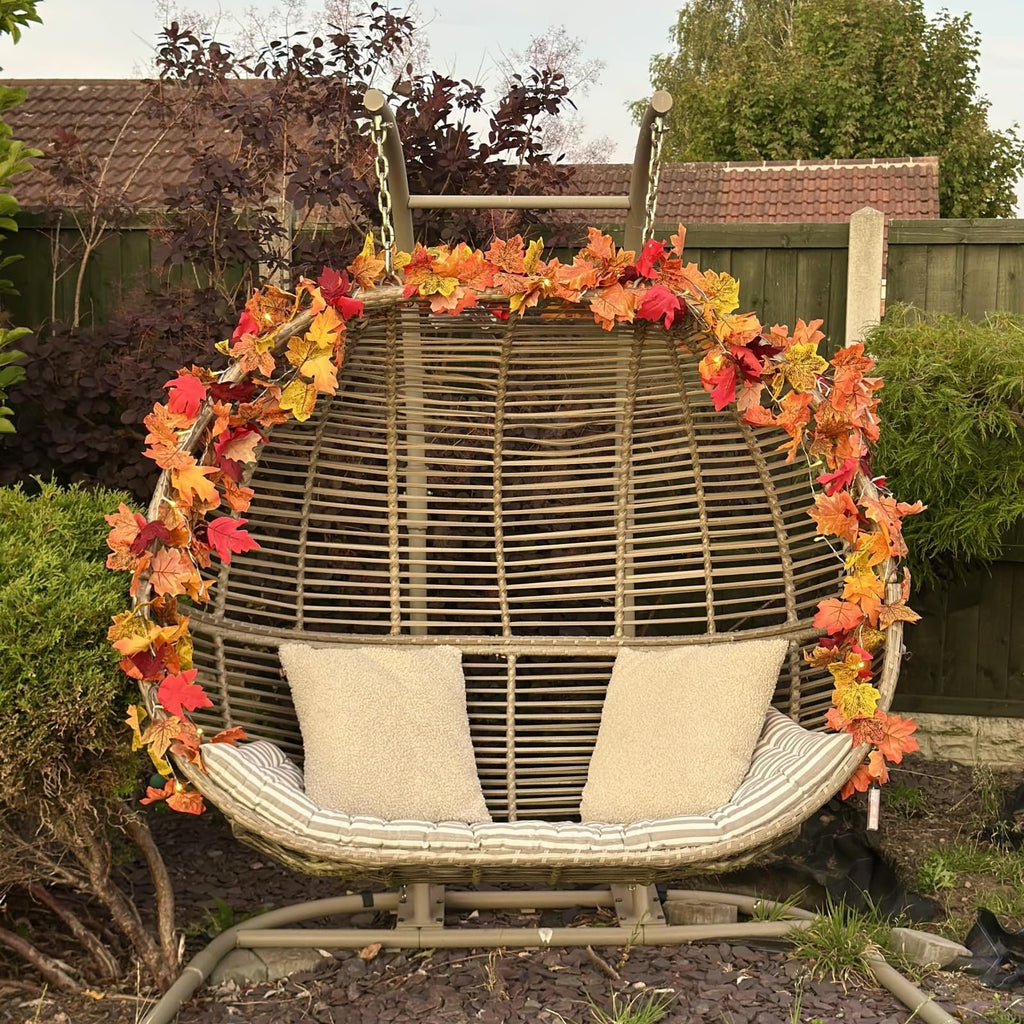 Artificial maple leaf garland with lights draped over a hanging egg chair with cushions in a garden setting.