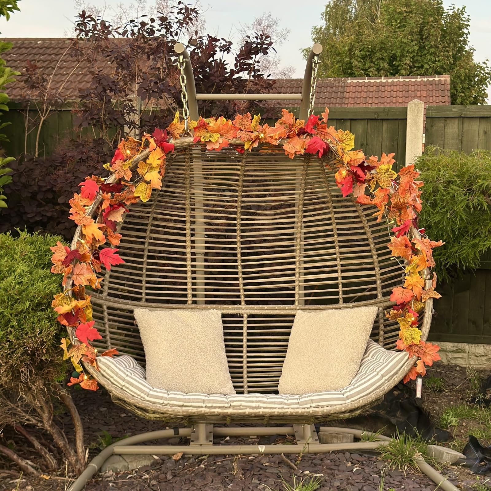 Artificial maple leaf garland with lights draped over a hanging egg chair with cushions in a garden setting.