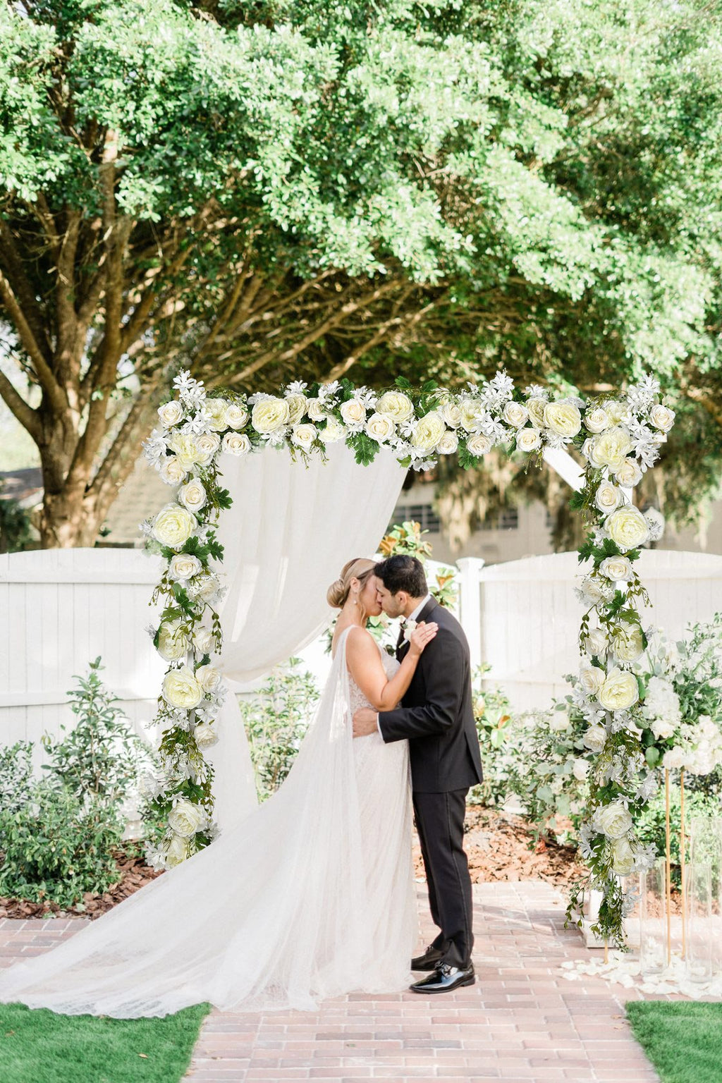 Wedding couple kissing under a floral arch decorated with white roses and greenery