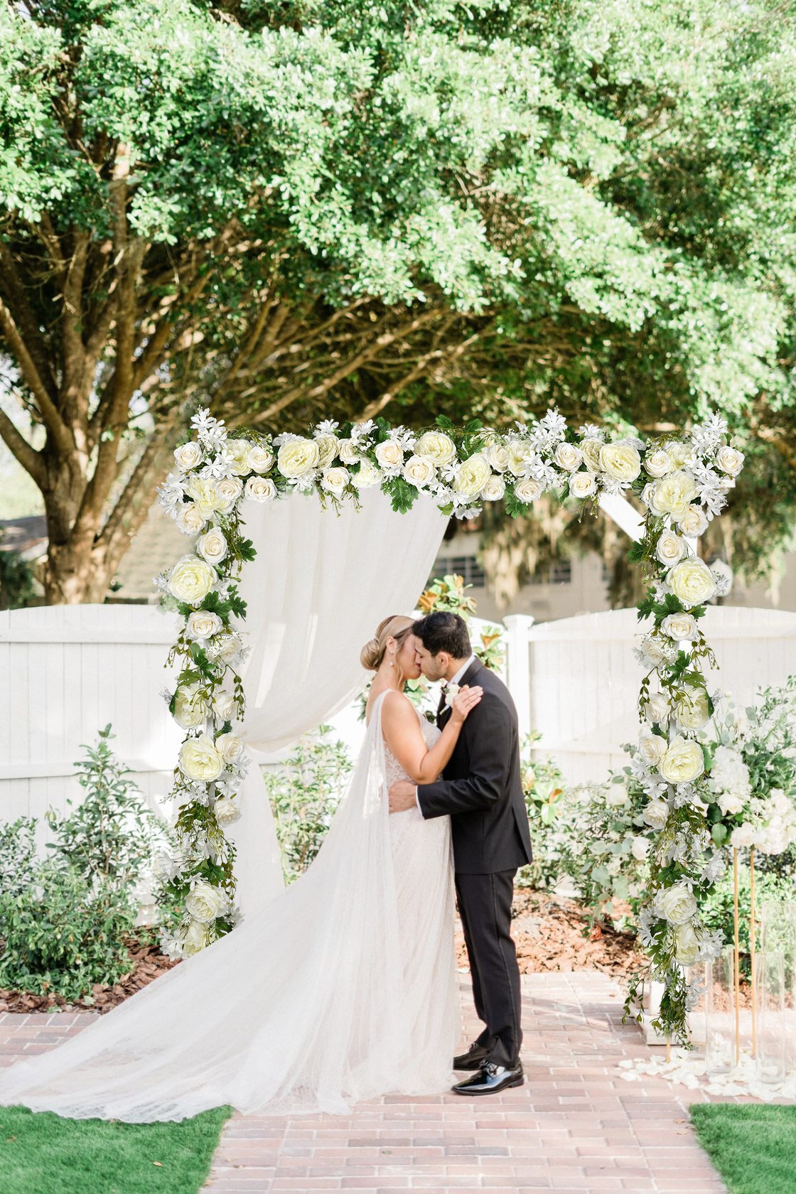 Wedding couple kissing under a floral arch decorated with white roses and greenery