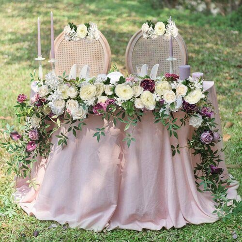 Wedding sweetheart table decorated with white and purple flowers and greenery, set outdoors on grass.