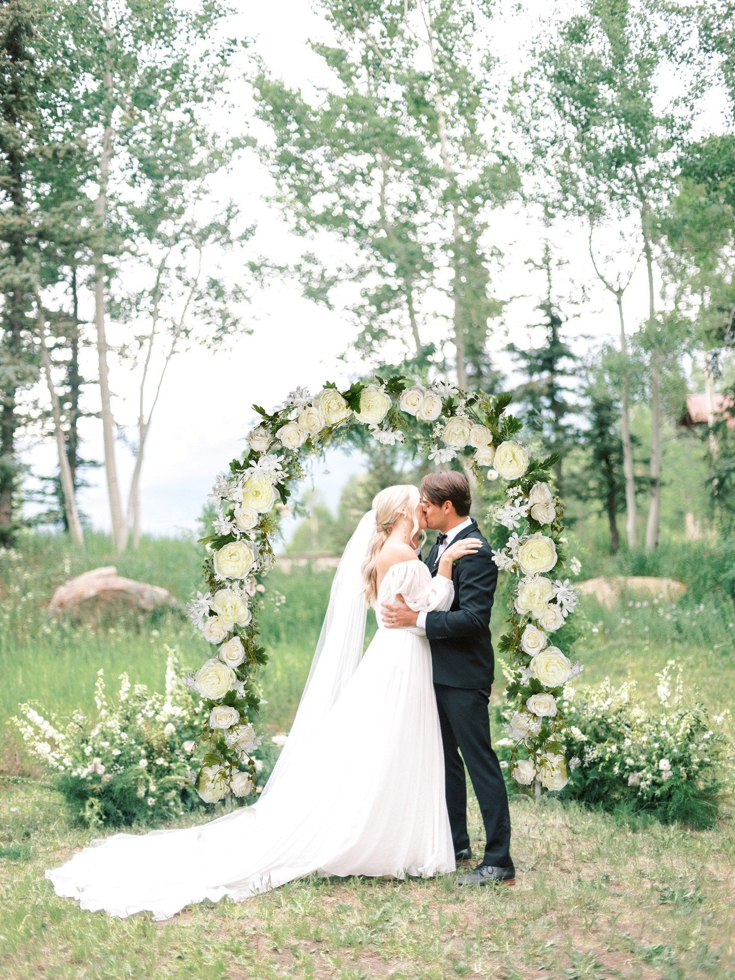 Bride and groom kissing under a floral wedding arch decorated with white roses and greenery