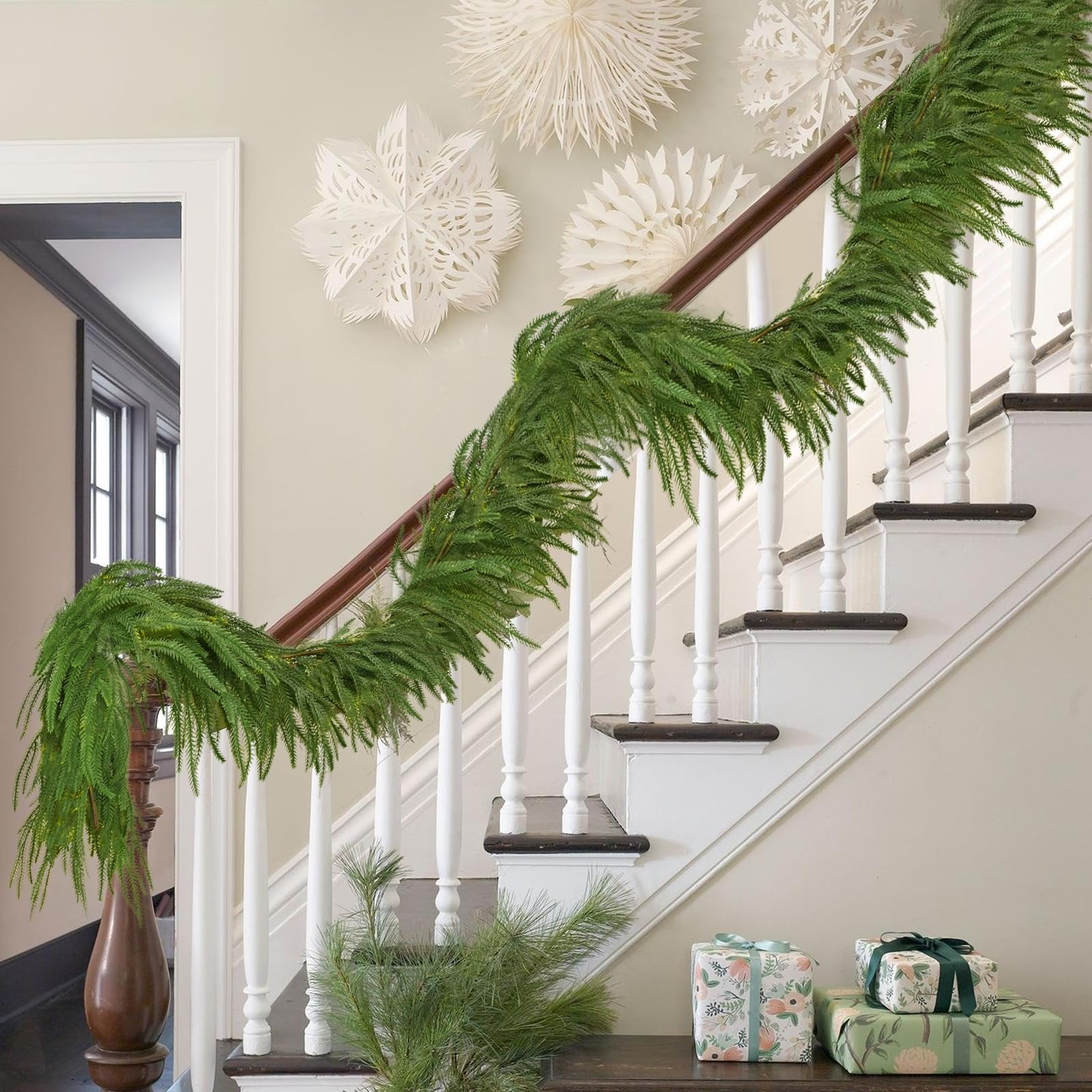 Green artificial Norfolk pine garland draped over a wooden banister on a white staircase, with decorative paper snowflakes on the wall and wrapped gifts below.