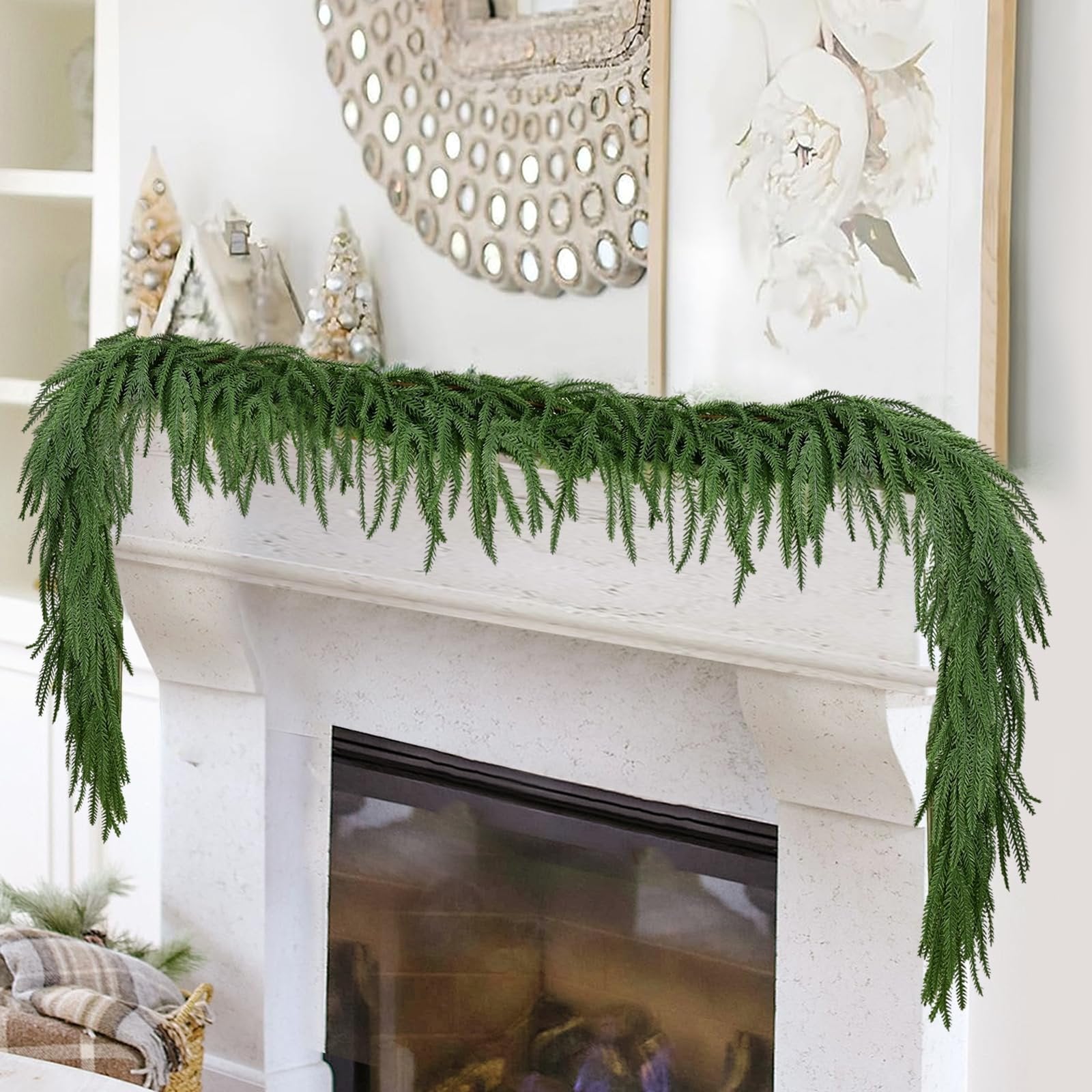 Green artificial Norfolk pine garland draped over a white fireplace mantel with decorative Christmas trees and artwork in the background.