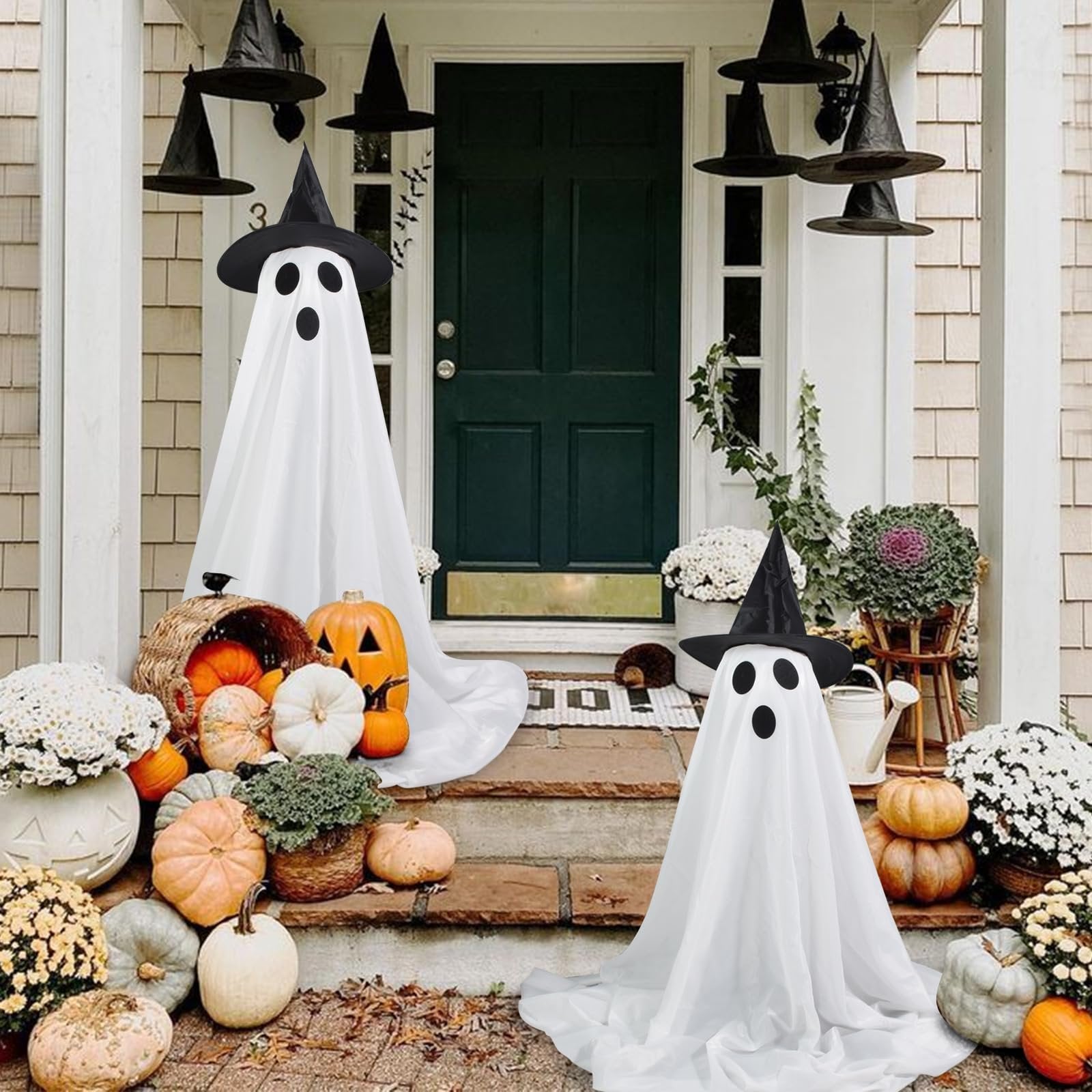 Two white fabric ghost decorations with witch hats and black faces stand on a porch with pumpkins and fall flowers.