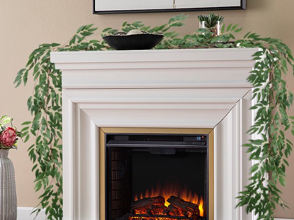 Artificial willow vine garland with green leaves draped over a white fireplace mantel with a decorative bowl and a vase with a flower.