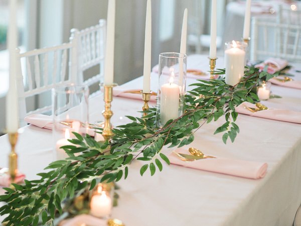 Artificial green willow vine garland draped across a white tablecloth with pink napkins and gold cutlery, surrounded by lit candles in brass and glass holders.