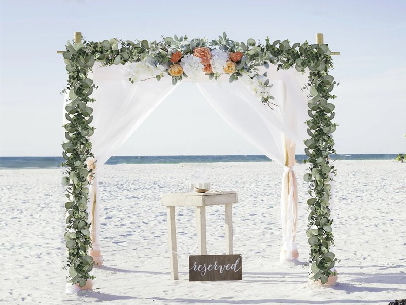 Wedding arch decorated with artificial eucalyptus garland and flowers on a beach.