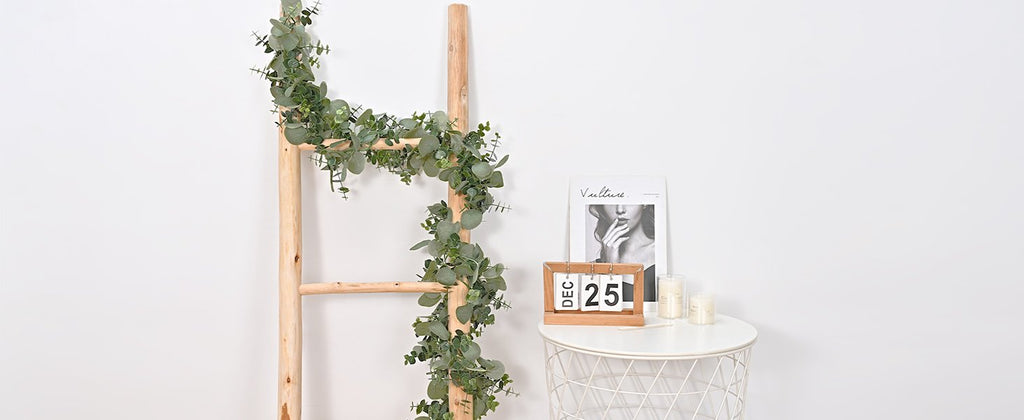 Artificial eucalyptus garland draped on a wooden ladder next to a small table with a perpetual calendar and candles.