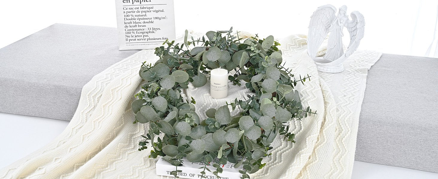 Artificial eucalyptus garland arranged in a circle on a book, with a candle and angel statue nearby.