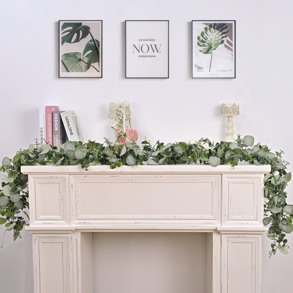 Artificial eucalyptus garland draped over a white mantelpiece with decorative items and framed art above.