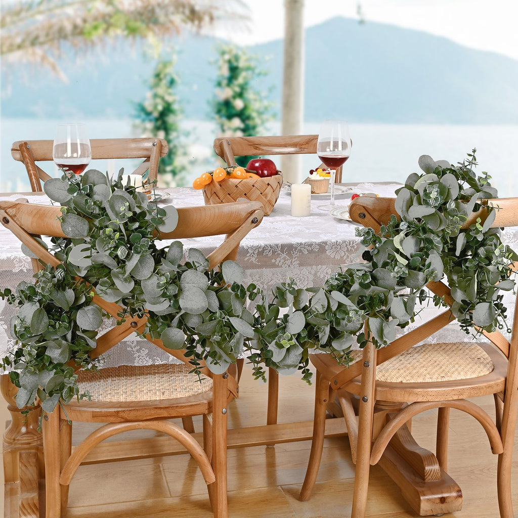 Artificial eucalyptus garland draped over wooden chairs at a table setting with wine glasses and fruit basket
