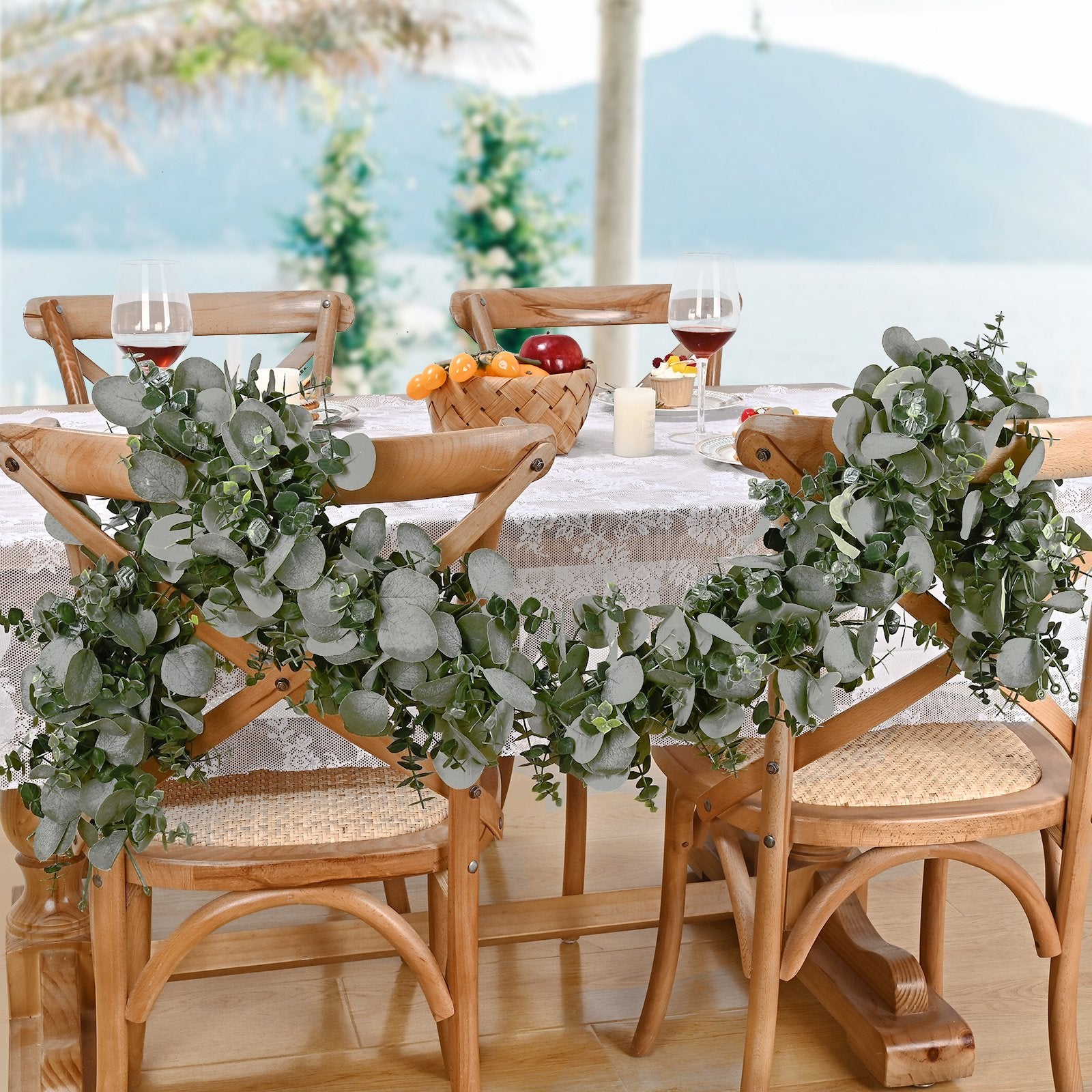 Artificial eucalyptus garland draped over wooden chairs at a table setting with wine glasses and fruit basket