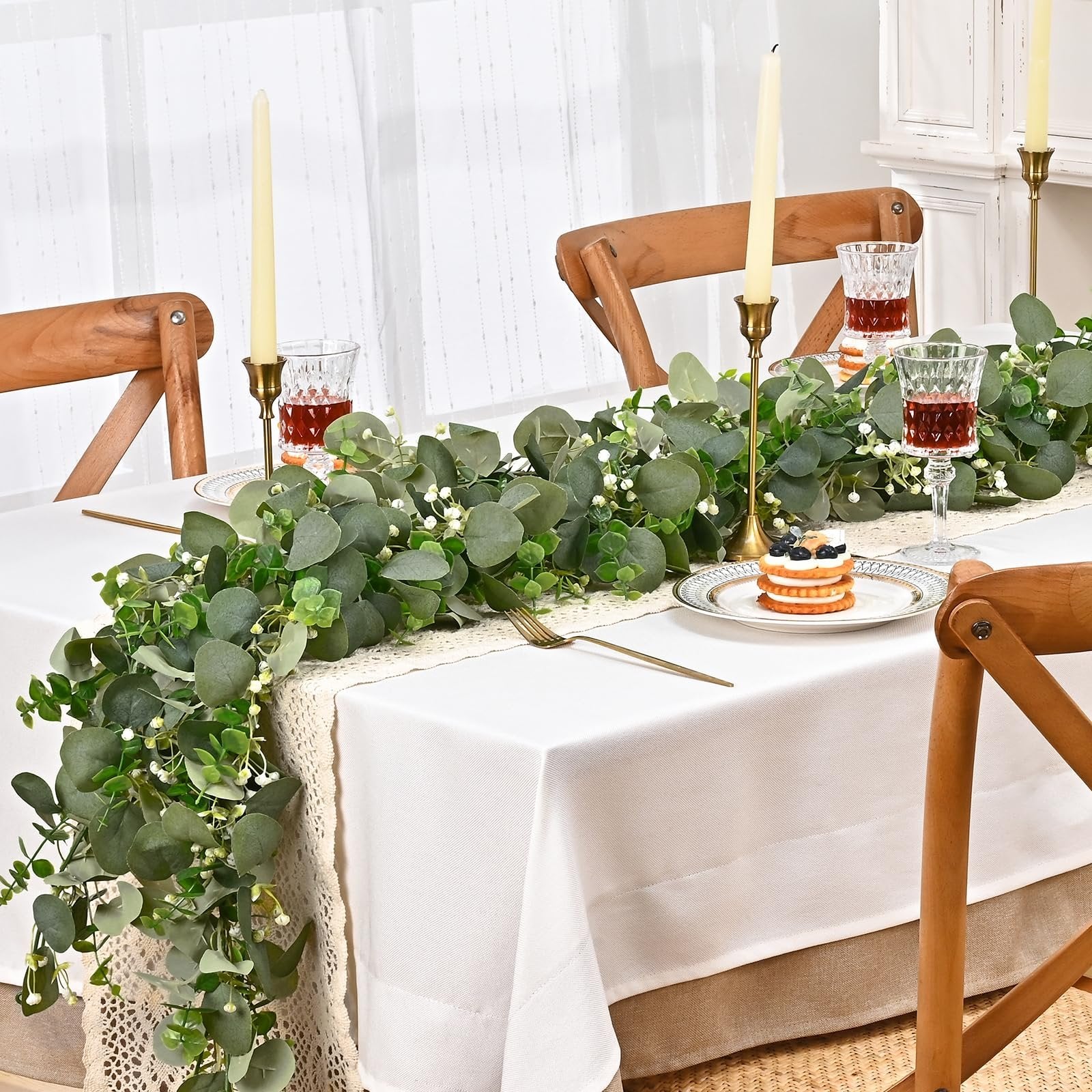 Artificial eucalyptus garland draped across a white tablecloth on a dining table with place settings.