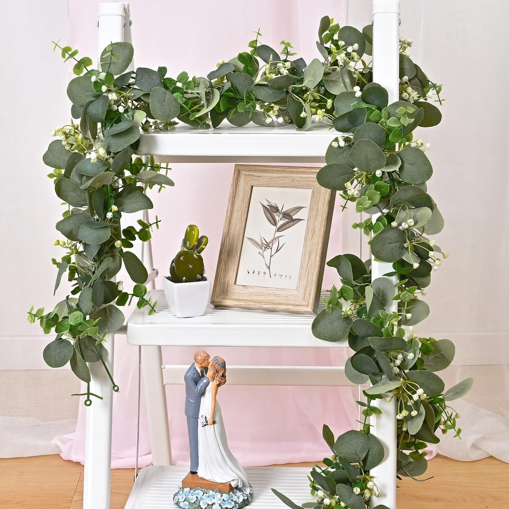 Artificial eucalyptus garland draped on a white shelf with a wedding couple figurine, a framed picture, and a small potted plant.