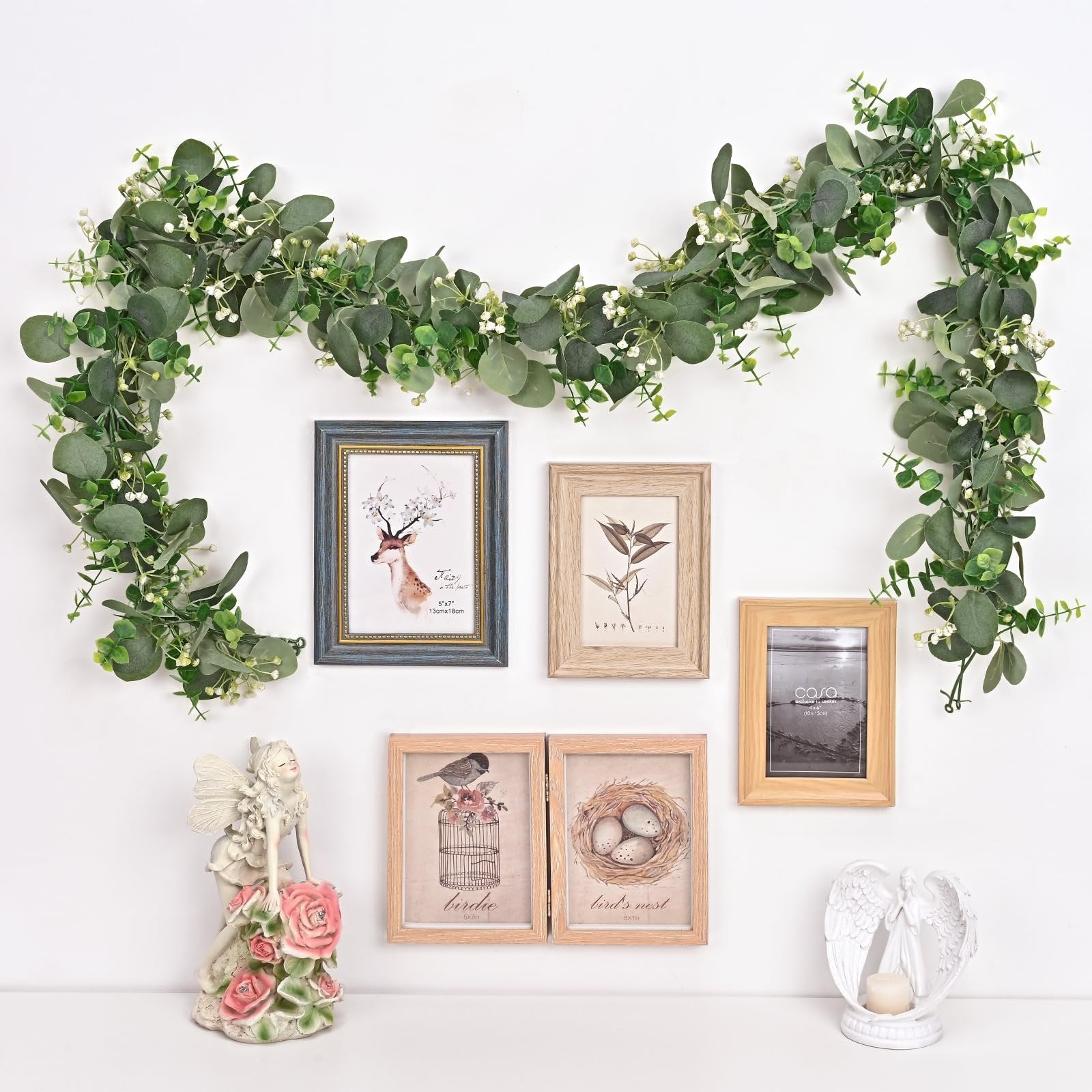 Artificial eucalyptus garland draped across a white wall with decorative picture frames and figurines.