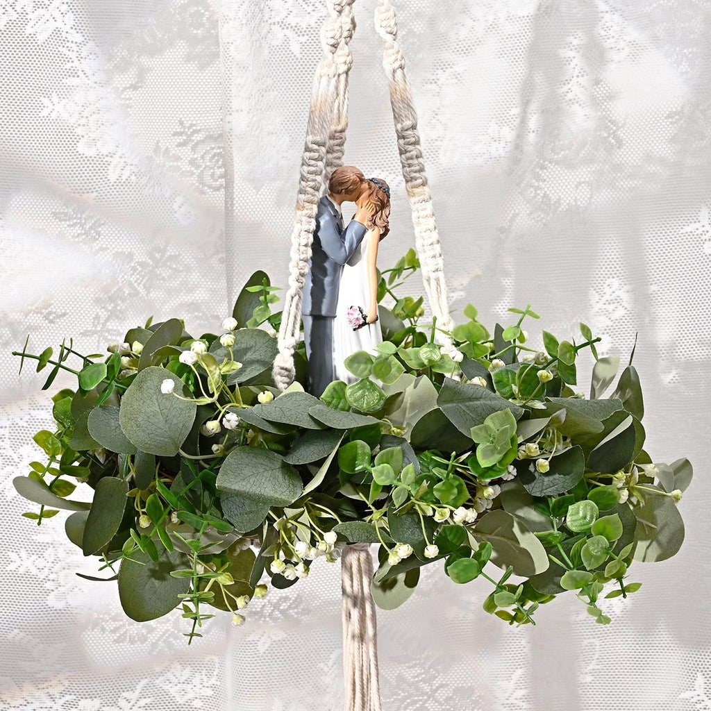 Wedding cake topper of couple kissing surrounded by artificial eucalyptus garland and greenery, hanging from macrame.