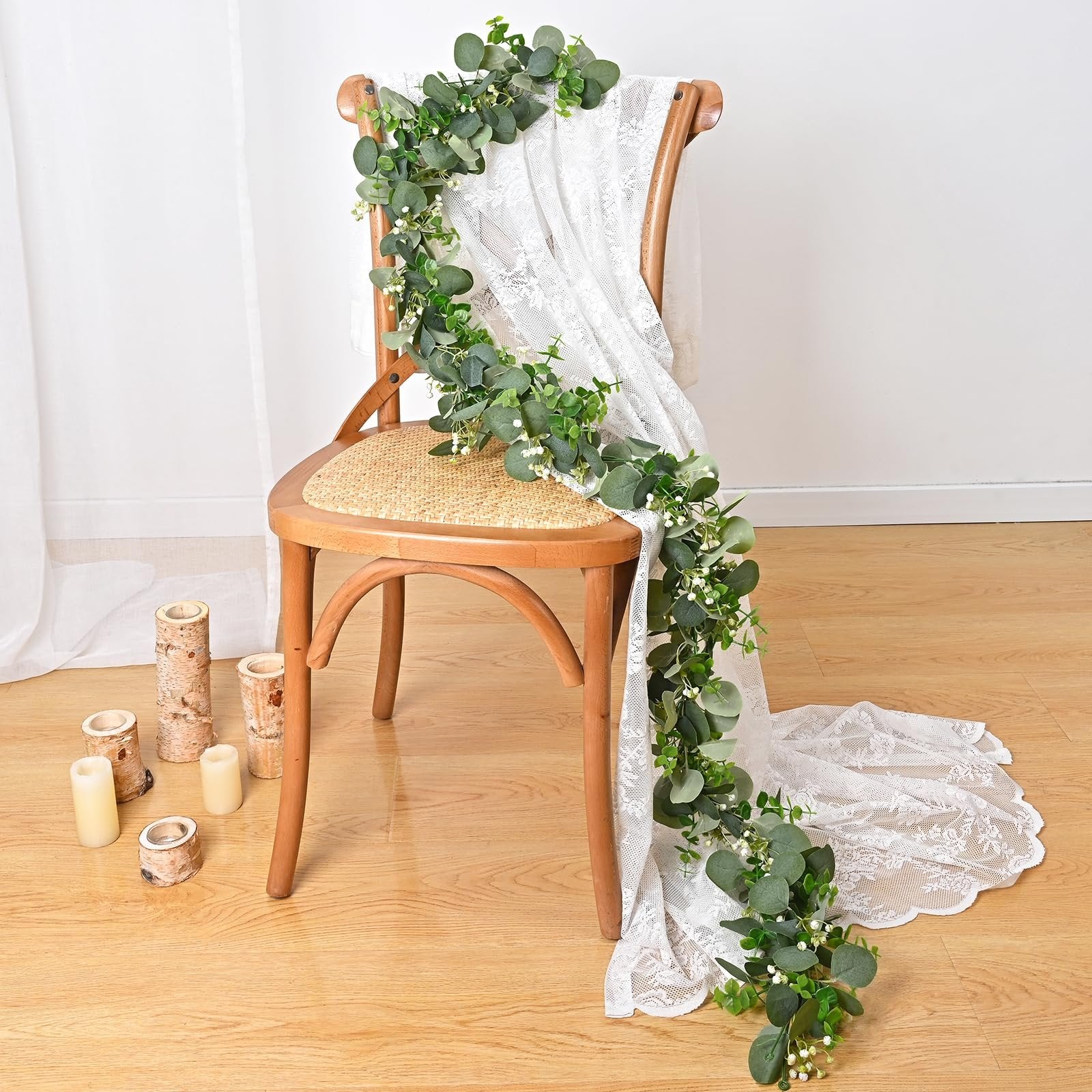 Artificial eucalyptus garland draped over a wooden chair with lace fabric and decorative logs and candles on the floor.