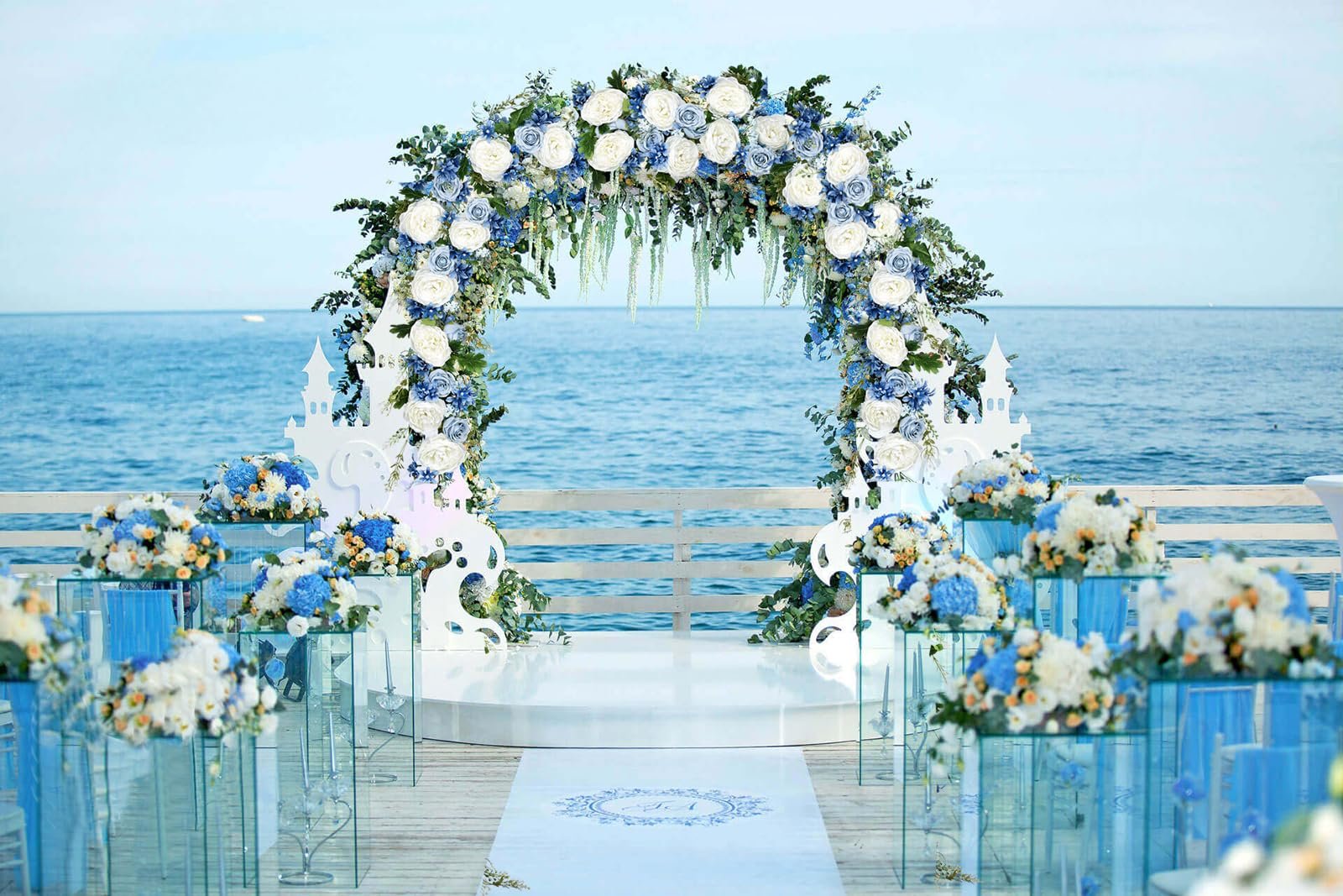 Wedding arch decorated with blue and white flowers, overlooking the ocean. A white aisle runner with a monogram is in the foreground.