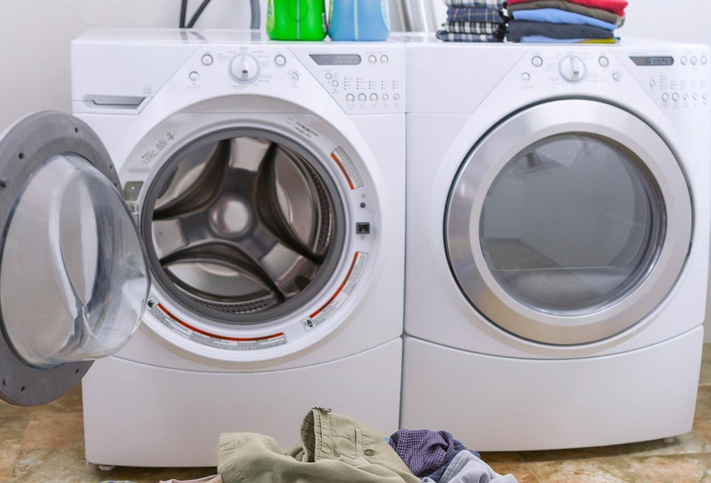 Front view of a white washing machine with the door open and a white dryer next to it, with laundry on the floor.