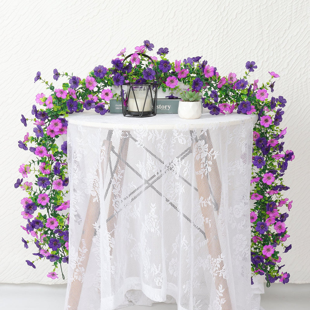 Artificial daisy flower garland with purple and pink blooms draped over a white lace-covered table.