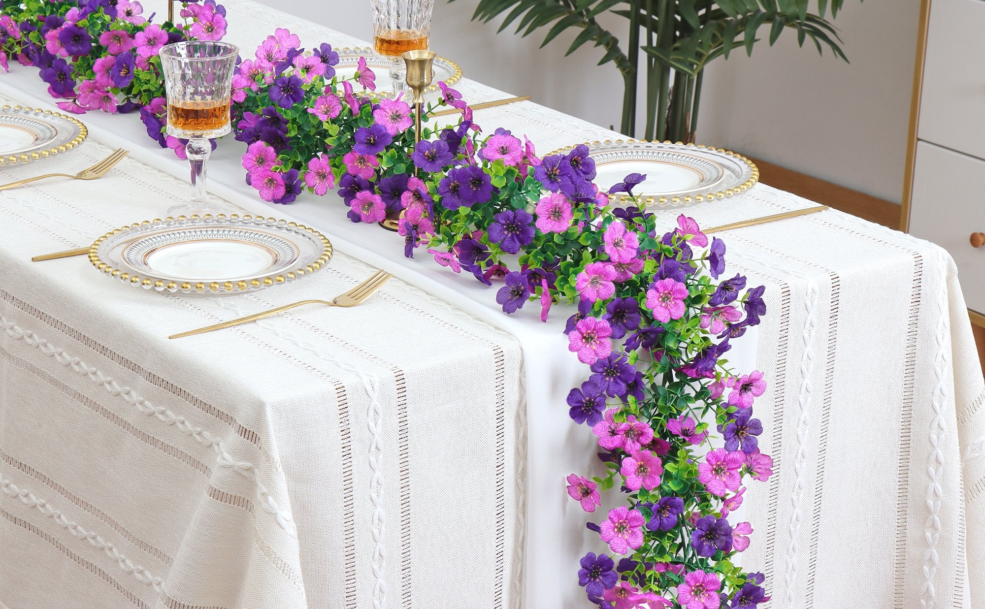 Artificial daisy flower garland in light dark purple draped across a white tablecloth with gold-rimmed plates and gold cutlery.