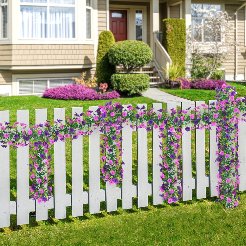 Two strands of artificial purple and pink daisy flower garlands draped over a white picket fence in front of a house.