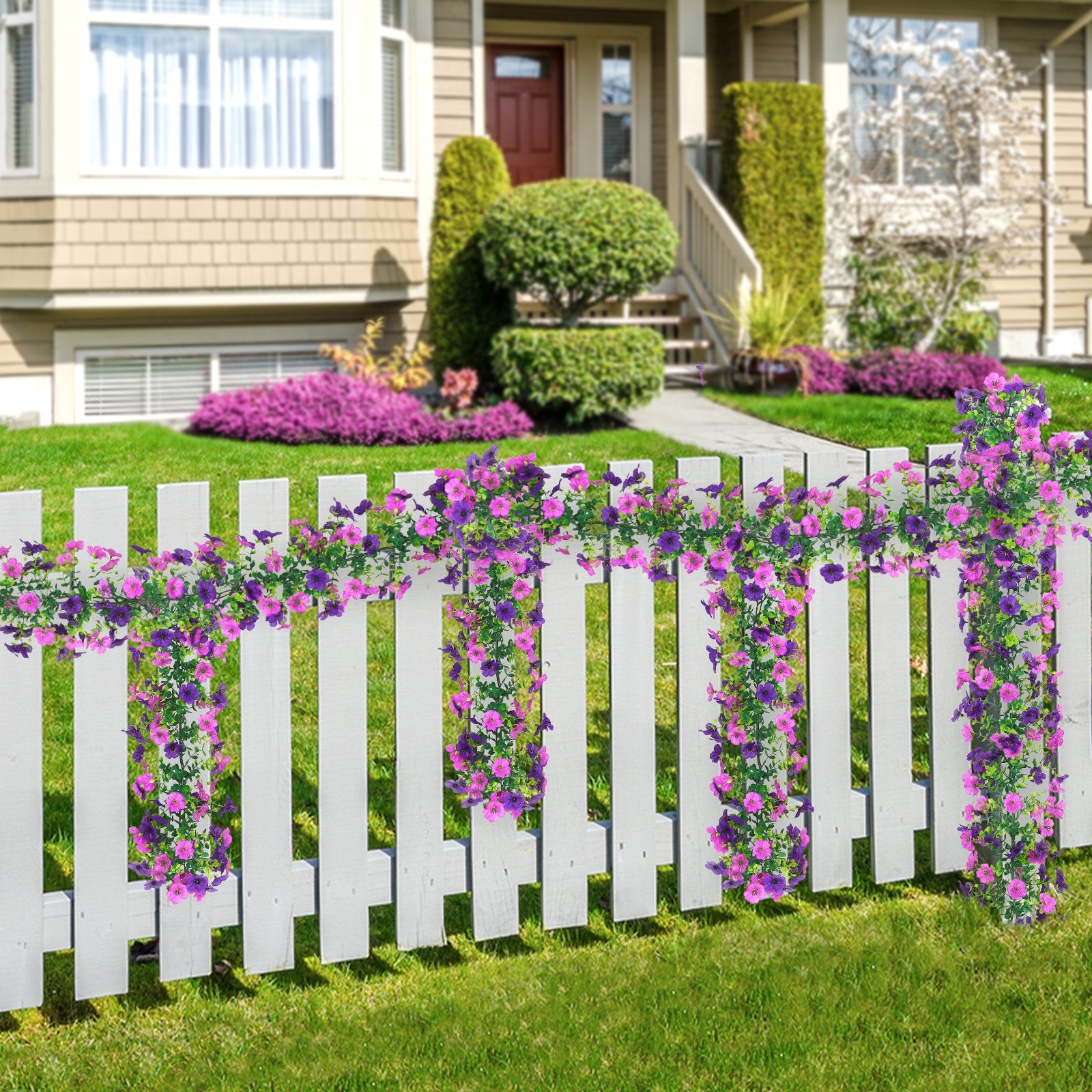 Two strands of artificial purple and pink daisy flower garlands draped over a white picket fence in front of a house.
