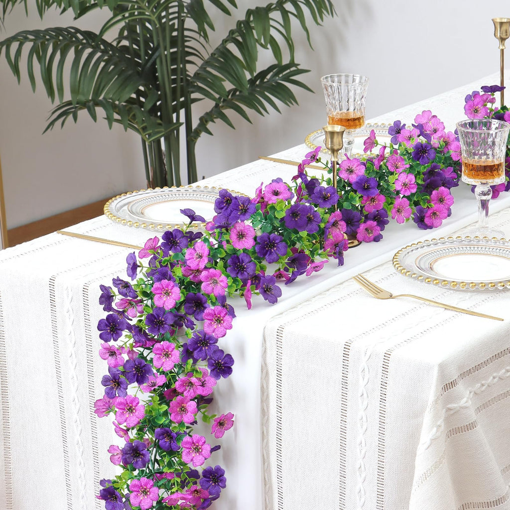Artificial purple and pink daisy flower garland draped across a white tablecloth with place settings and glasses.