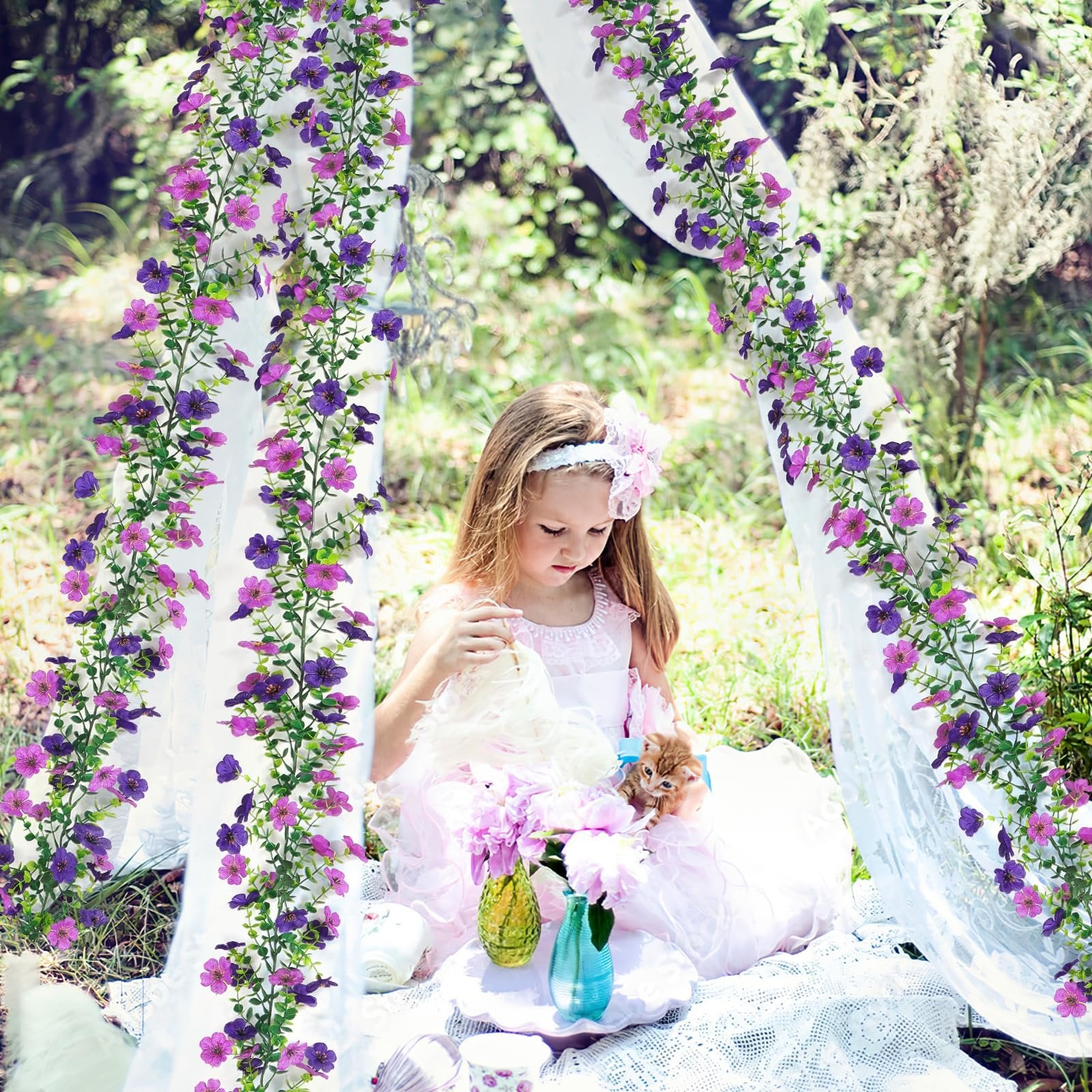 Young girl in a pink dress holding a kitten and a doll, seated outdoors under floral garlands.