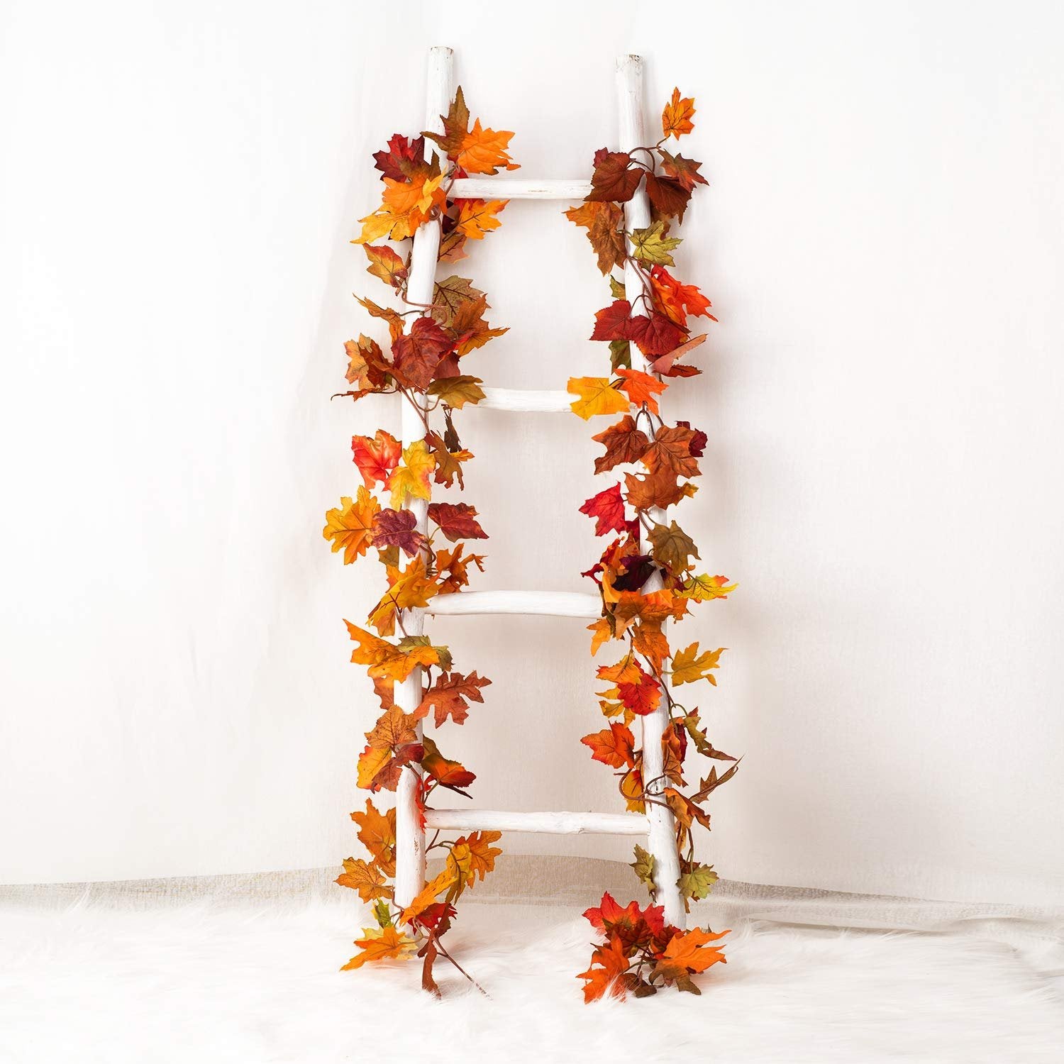 Two artificial fall maple leaf garlands draped on a white wooden ladder against a white backdrop.