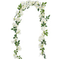 Artificial gardenia garland with white silk flowers and green leaves on a white background, shown draped in an arch shape.