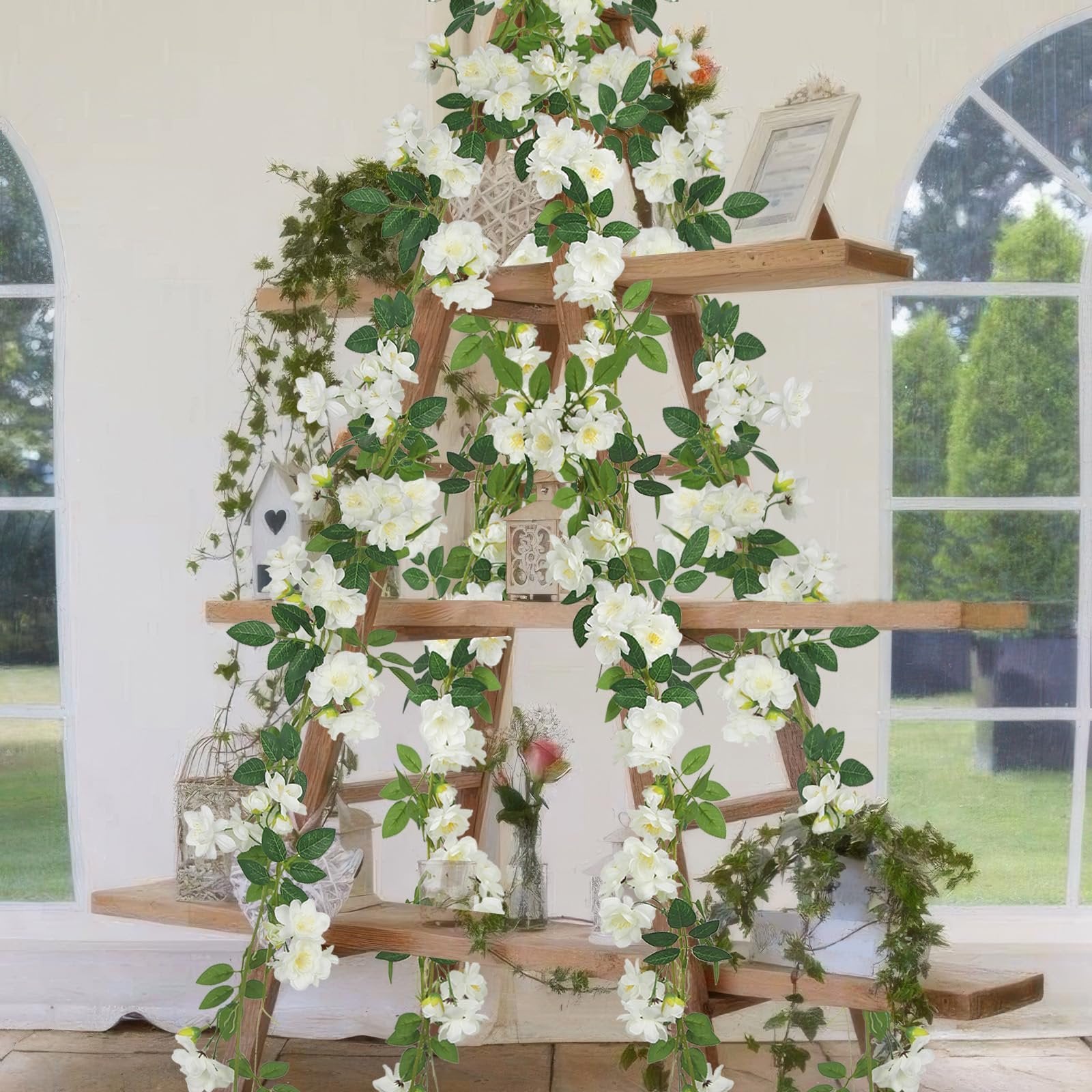 Artificial gardenia garland with white flowers and green leaves draped on a wooden ladder shelf with decorative items.