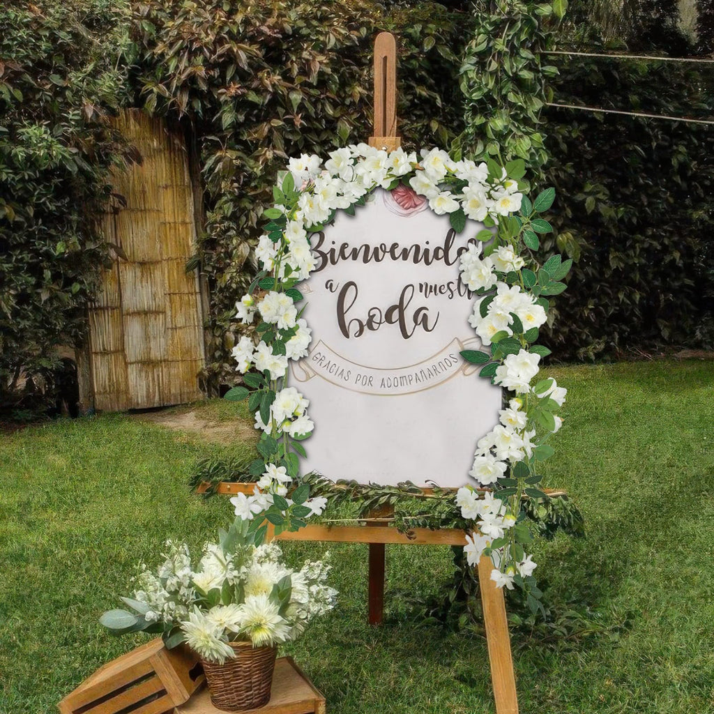 Wedding welcome sign decorated with white flowers and greenery, with a floral arrangement below.