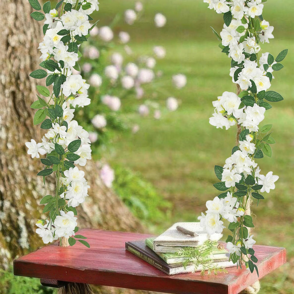 Two artificial gardenia garlands with white flowers and green leaves draped around a tree trunk and over a red table with books.