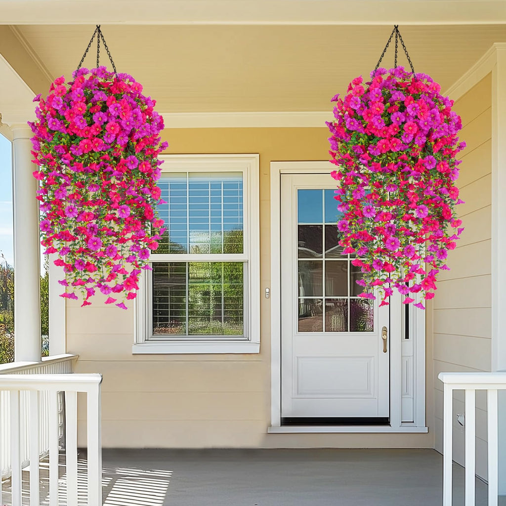 Two hanging baskets of artificial fuchsia and rose red petunia flowers adorn a porch with a window and door.