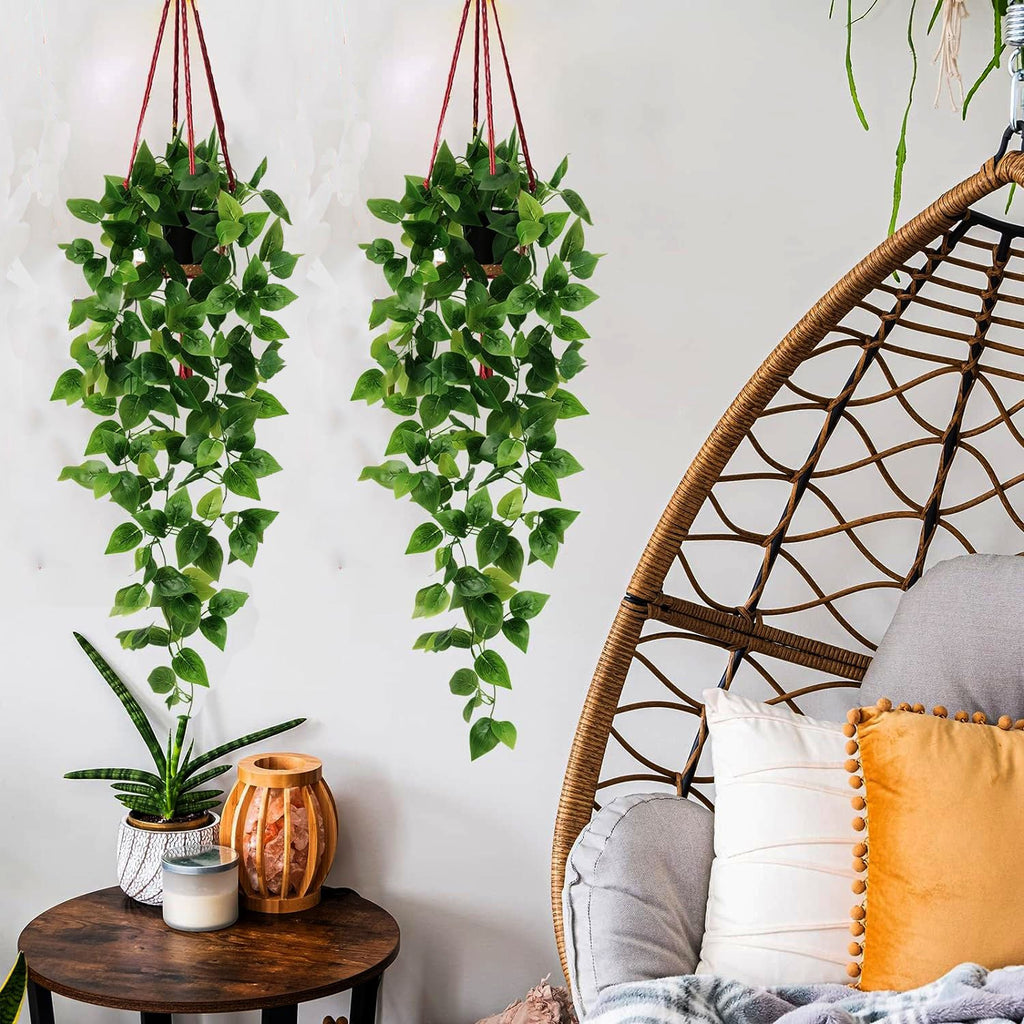 Two artificial hanging ivy plants with black pots suspended on red ropes, displayed in a room with a rattan chair and side table.